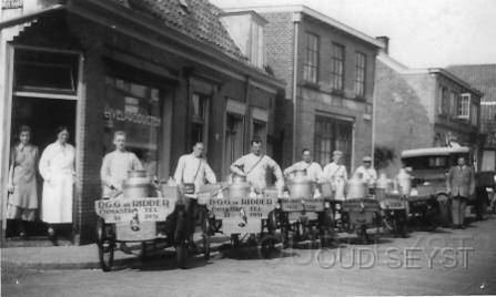 Emmastraat-1920-001.jpg - Foto van de Melkhandel De Ridder Emmalaan 21 te Zeist. De melkventers (van links naar rechts): Bob van Donselaar, Jan van Hall, Gijs van Hall, Ko van Hall, Frans van Donselaar,(broer van Bob) en Henk van Delft. De baas Dirk de Ridder staat bij de Ford. In de deuropening rechts de bazin Ali de Ridder en links Aaltje van Veluw (latere schoonzus van Bob van Donselaar). De melkhandel van D.G.G. de Ridder was gevestigd aan de Emmastraat (Achterheuvel) no: 2. Deze melkhandel was hier gevestigd vanaf omstreeks 1929 tot 1938. In 1939 kwam hier J. de Haan en in 1963 J. van de Munt in deze melkhandel. Opname van 1937.